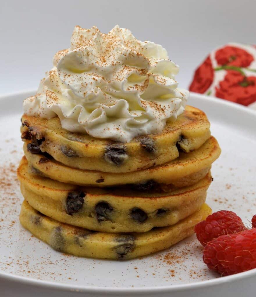 A plated stack of Cannoli Pancakes topped with whipped cream and raspberries on the side